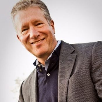 A man with short gray hair wearing a blue shirt, blue sweater, and gray suit jacket smiles for the camera.