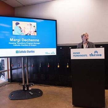 A woman talks into a microphone behind a podium, next to a large screen with slides.