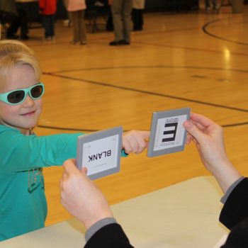 A young girl wearing a teal-colored shirt and green glasses points at one of two cards that a woman holds up inside a school gym.