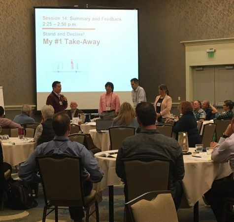 A group of adults gathered around circular tables look up at two speakers, who stand in the front of the room with slides that say