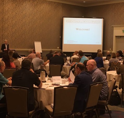 A room full of people sitting around circular tables with a projector showing a slide that says "Welcome!" in the background.