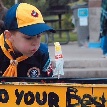 A boy in a blue and yellow hat blows at something on a railing.