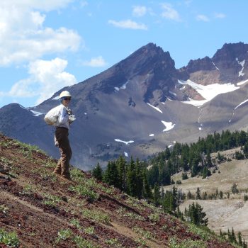 A person wearing a white hard hat smiles at the camera while on a hill with mountains in the background.