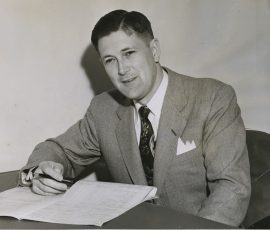 A black and white photo of Jack Murdock, a man with short dark hair wearing a gray suit writing something on a piece of paper