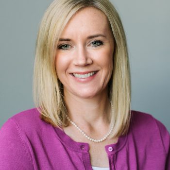 A woman with straight blond hair wearing a purple sweater and a string of pearls smiles for the camera.