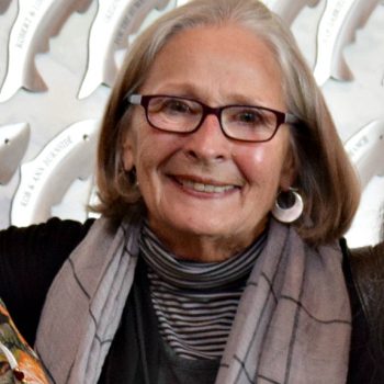 A woman with short gray hair wearing a gray scarf, silver earrings, and glasses smiles for the camera.