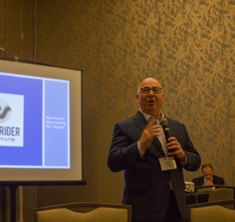 A man wearing a suit and glasses speaks into a microphone with presentation slides on a screen behind him.