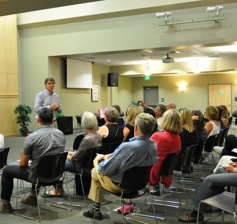 A man with gray hair speaks to an audience of seated adults inside a room.