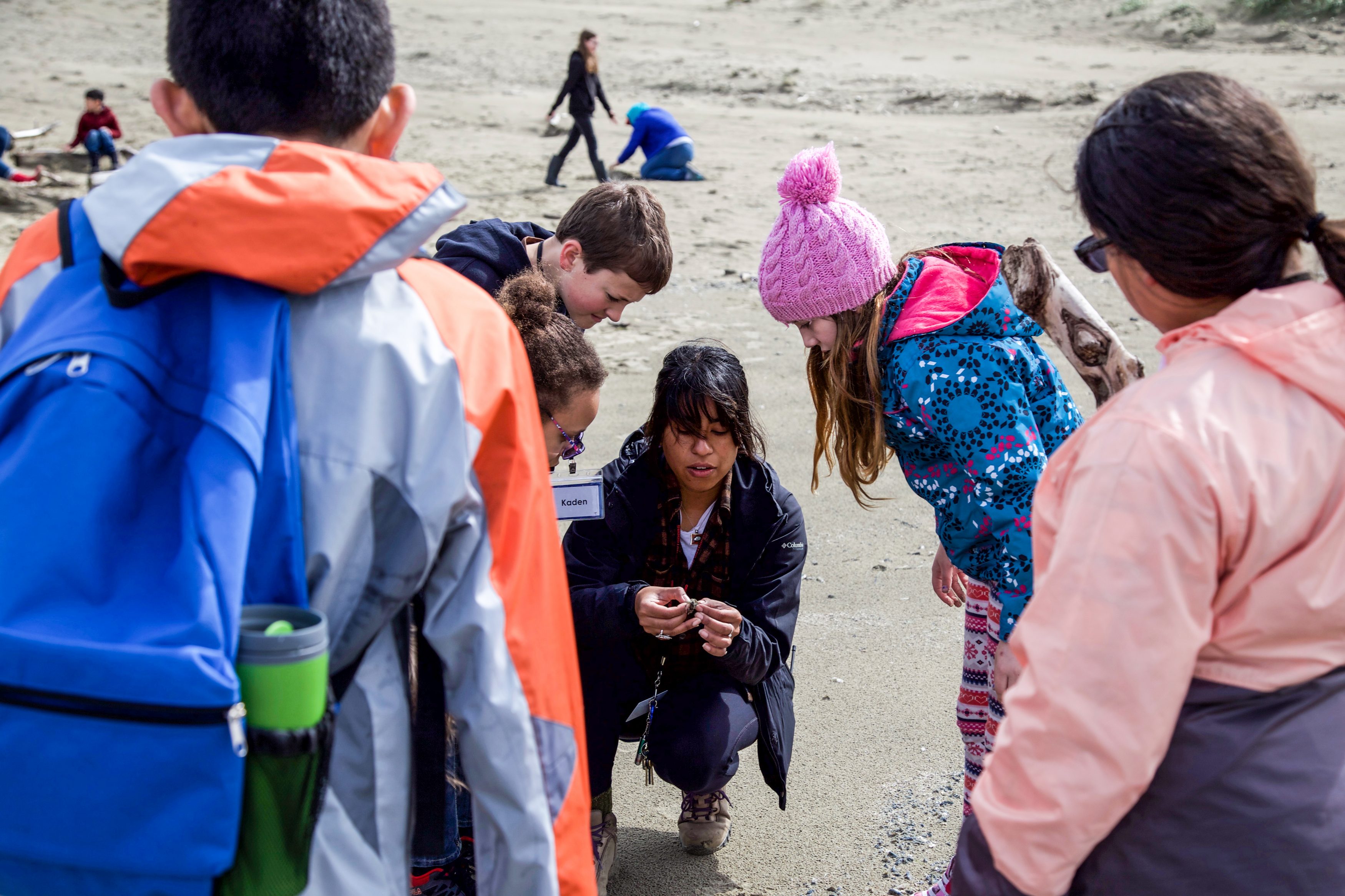 A woman with dark straight hair holds something in her hand, while children gather around her to observe on the beach.