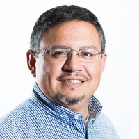 A man with dark gray hair wearing glasses and a blue checkered shirt smiles for the camera.