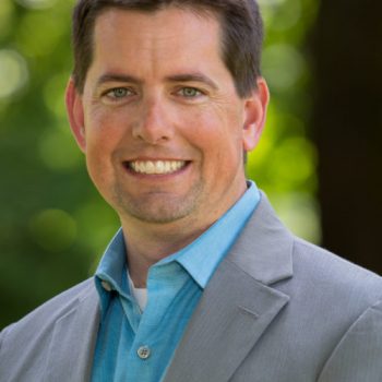 A man with short dark hair wearing a blue shirt and gray suit smiles for the camera outside.