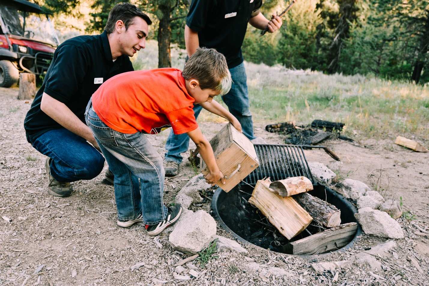 A young boy wearing an orange t-shirt and jeans dumps wooden logs onto a firepit while an adult wearing a black t-shirt helps.