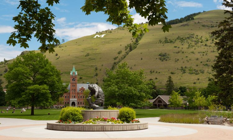 A college campus with red brick buildings and a fountain in front of a large green hill.