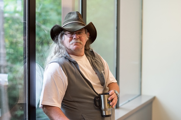 A man with long, straight gray hair and a mustache wearing a green cowboy hat, white t-shirt, and gray vest holding a coffee thermos looks at the camera near a window.