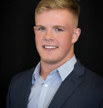 A young man with blond hair wearing a blue shirt and suit jacket smiles at the camera.