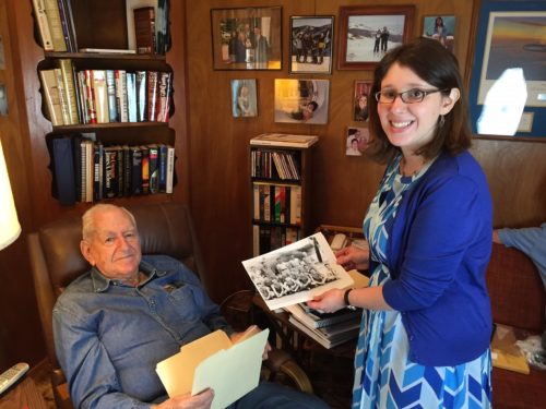 A woman with short dark hair wearing glasses, a blue dress, and a blue sweater holds a photo near a man with white hair and a mustache wearing a blue shirt sitting in a chair. They are in a living room with a bookshelf and photos on the wall behind them.