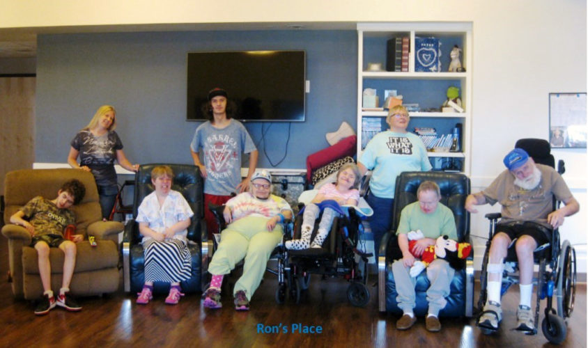 A group of people in wheelchairs or sitting in armchairs look at the camera with a TV and bookshelves behind them.