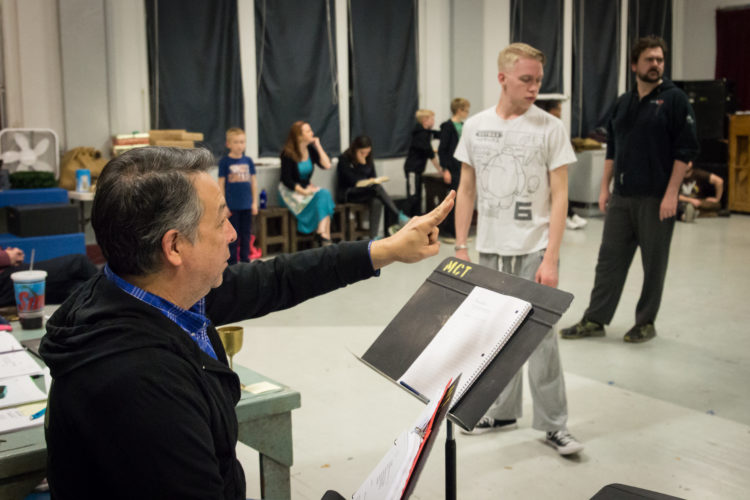 A man wearing a blue shirt and black jacket instructs actors in a studio.