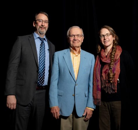 A man with gray facial hair wearing glasses, a blue shirt, and a gray suit, a man with gray hair wearing glasses and a blue suit, and a woman with long brown hair in a braid wearing a red scarf smile for the camera in front of a black background.