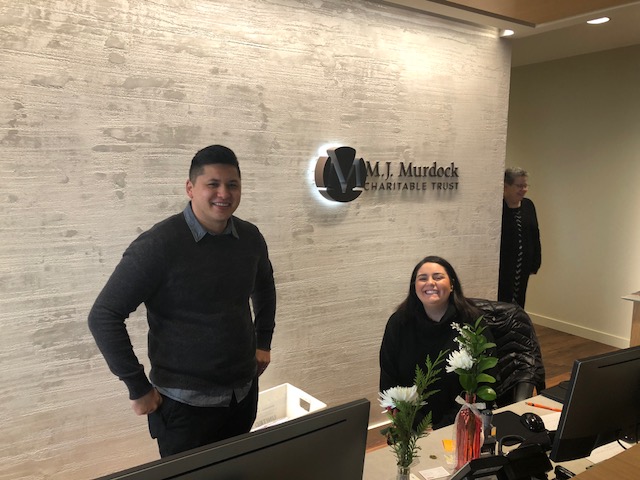 A man with short dark hair wearing a gray sweater stands next to a girl with straight black hair sitting at a front desk. The words "M.J. Murdock Charitable Trust" are on the wall behind them.