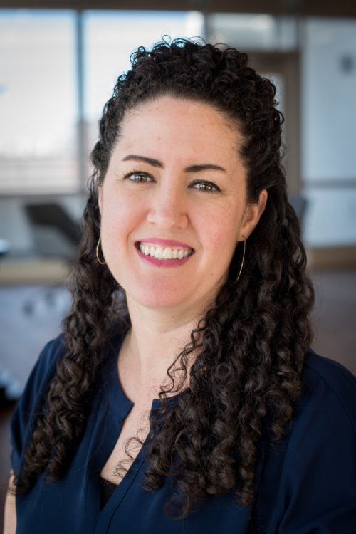 A woman with long, dark, curly hair wearing gold hoop earrings and a blue shirt smiles for the camera.