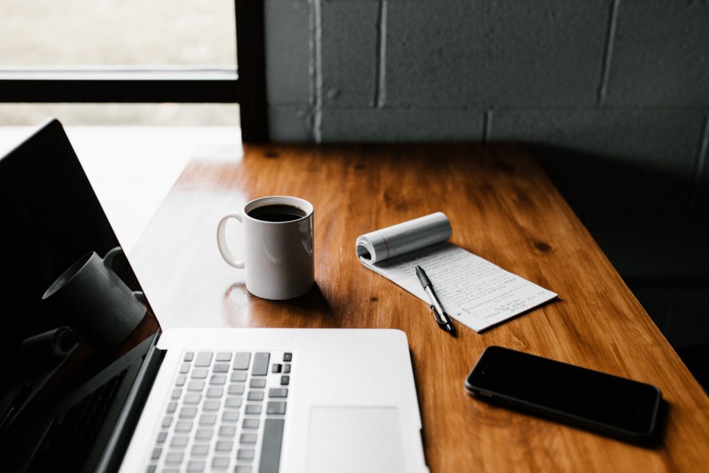 A wooden table with a laptop, cup of coffee, notebook, pen, and iPhone on it.