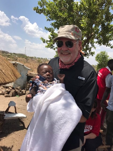 A man with white facial hair wearing sunglasses and a baseball cap smiles at the camera while holding a young baby wrapped in a white blanket while standing in a Uganda refugee camp.