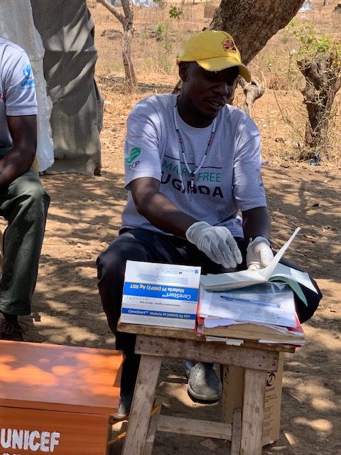 A man wearing a yellow baseball cap sits in front of a small wooden table at a refugee camp in Uganda.