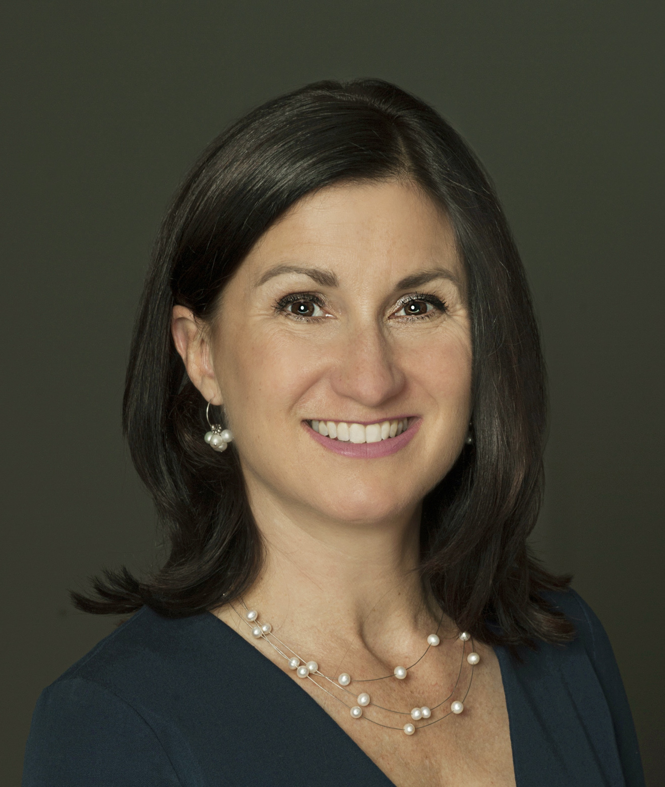 A woman with straight dark shoulder-length hair wearing pearl earrings, a pearl necklace, and a dark shirt smiles at the camera.