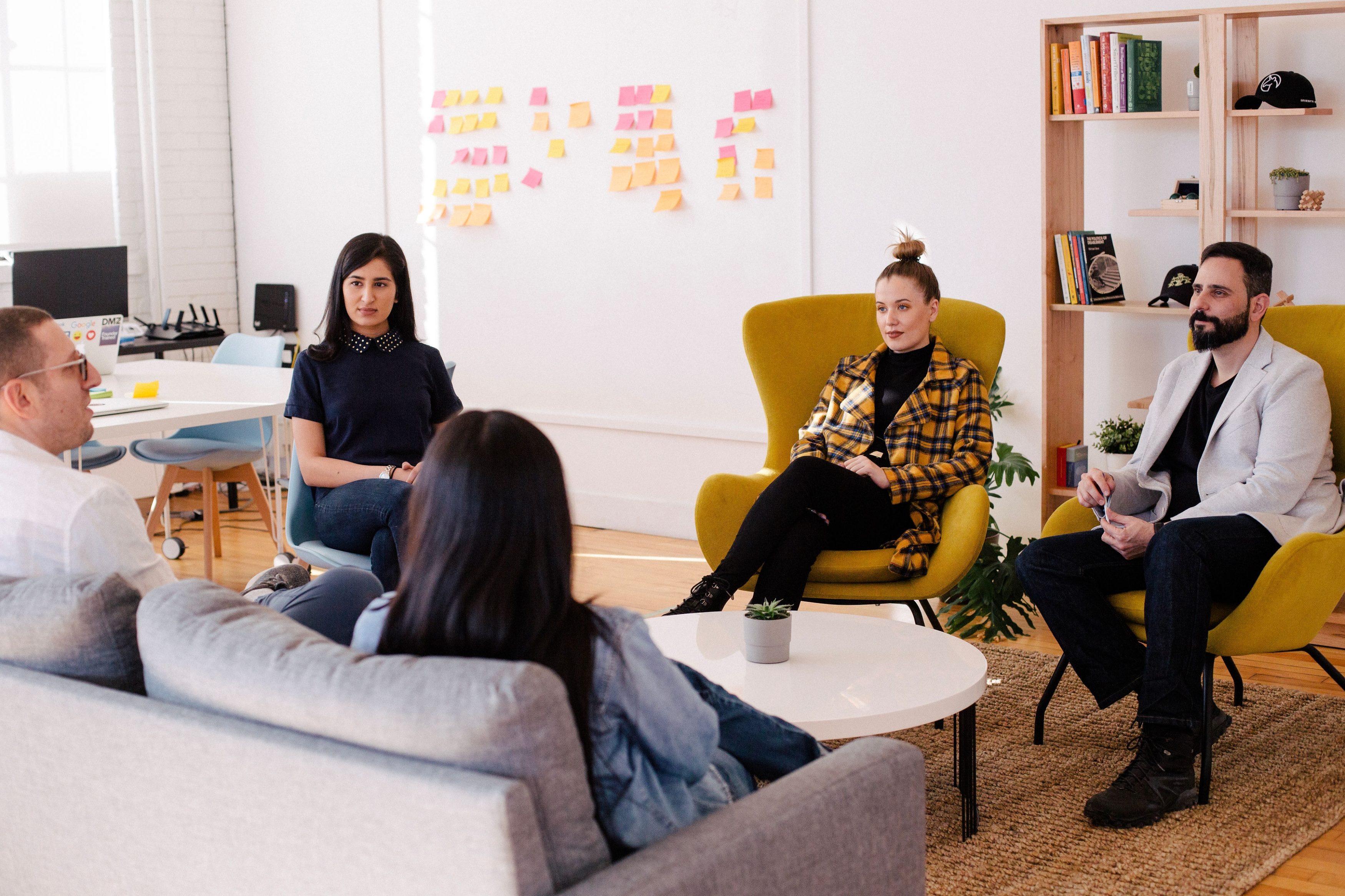 Five people sit inside a collaborative work space with a bookshelf, a wall with sticky notes, and a white table with computers in the background.