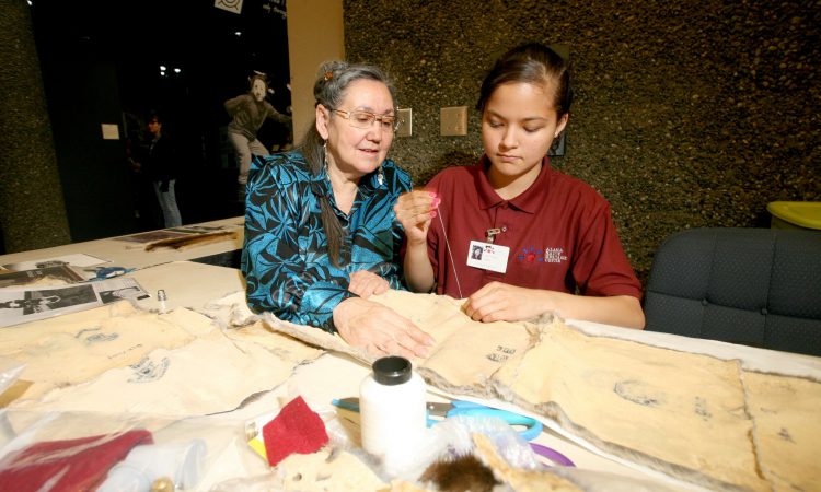 A woman with gray hair and glasses wearing a black and blue floral shirt helps a young girl with dark hair wearing a red shirt as she stitches onto fabric.