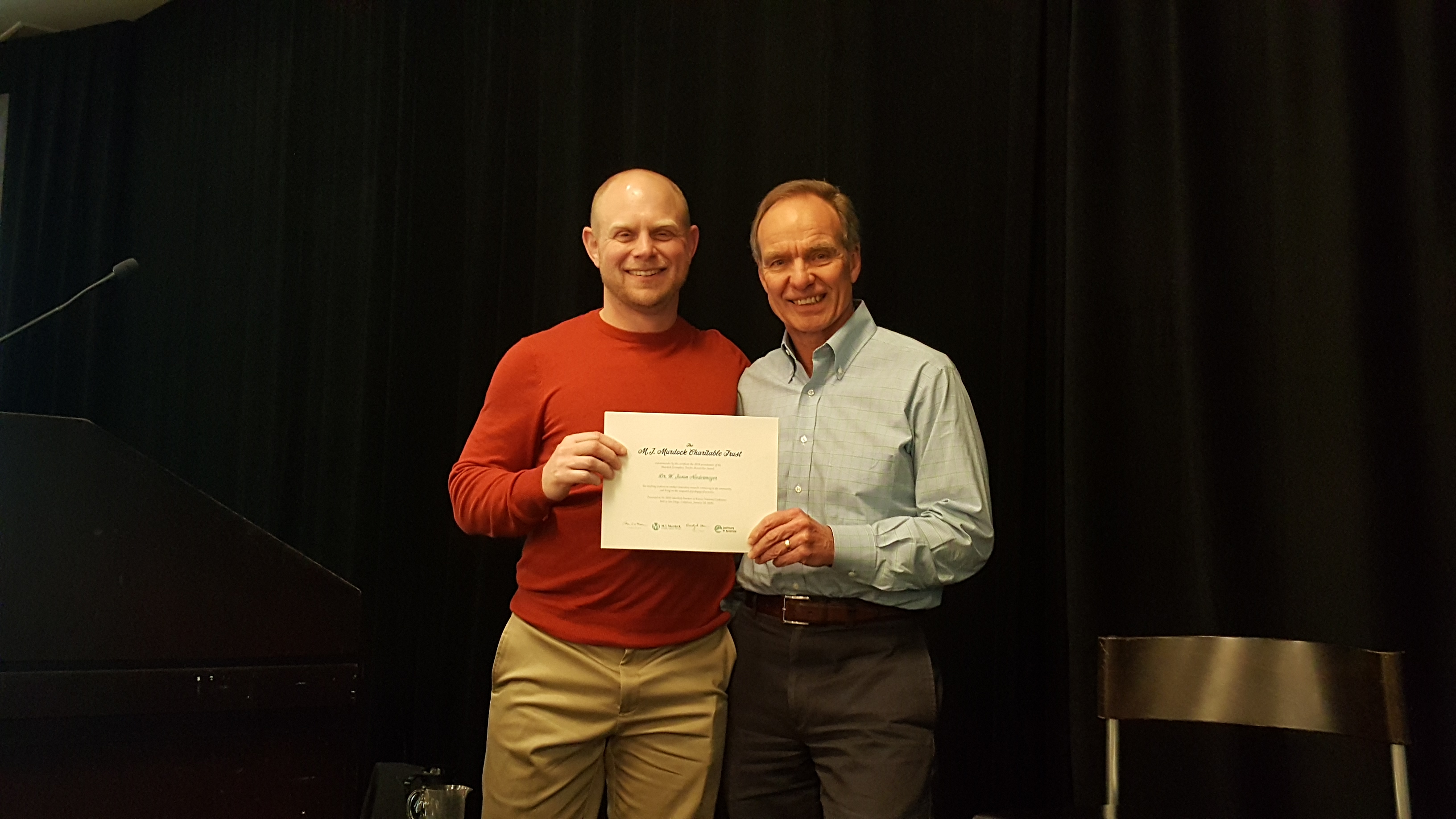 A man wearing a red shirt and a man wearing a blue button-up shirt smile for the camera while holding a certificate.