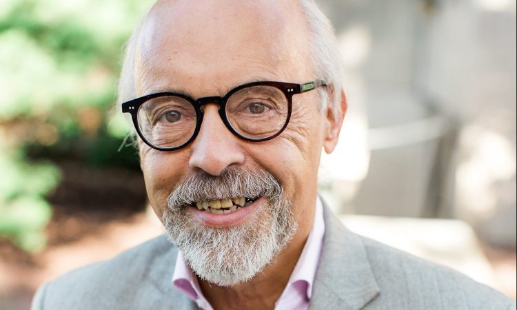 A man with white hair and black glasses smiles at the camera while standing outside.