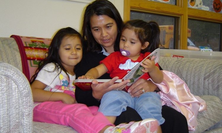 A woman with dark hair reads with two young children with dark hair while sitting on a couch inside.