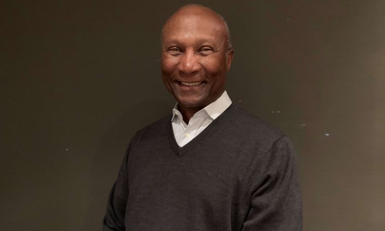 A man wearing a white shirt and gray sweater smiles at the camera.