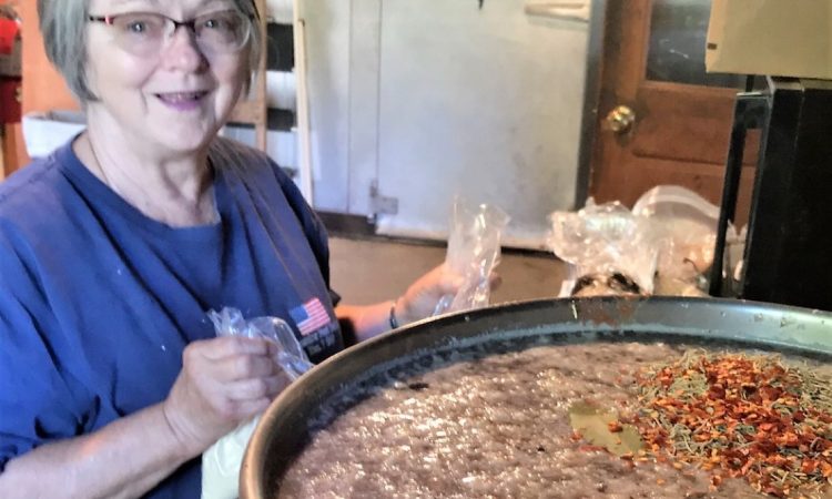 A woman with short gray hair and glasses wearing a blue t-shirt stands next to a large pot of food.