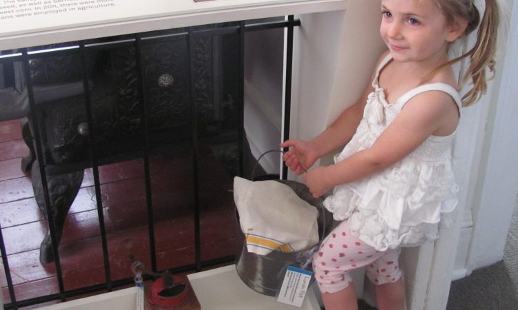 A young girl wearing a white shirt and pink leggings holds a tin pail next to a museum exhibit.