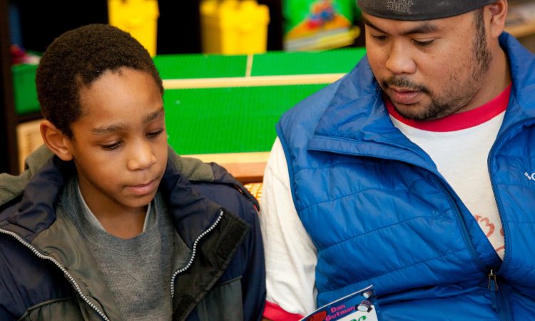 A young boy with black hair wearing a green and blue jacket reads a book with a man with facial hair wearing a blue vest and white shirt.