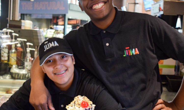 A young man wearing a black shirt puts his arm around a younger boy wearing a black shirt and black hat holding an ice cream sundae.