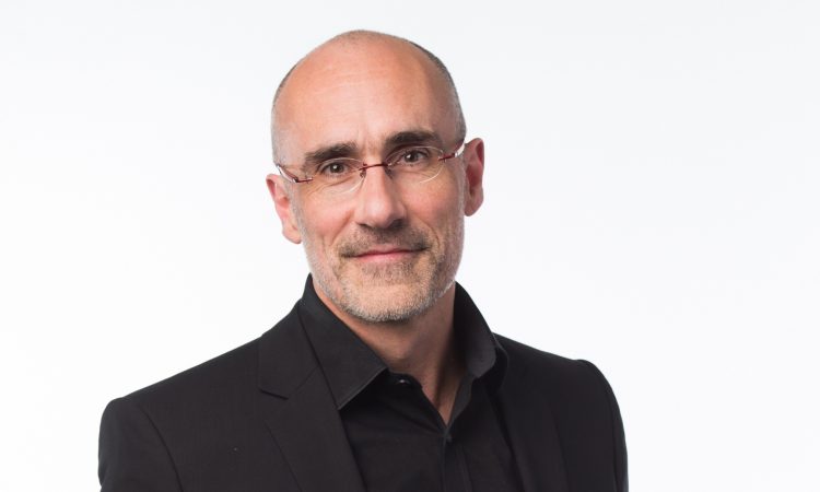 A man wearing glasses and a black suit looks at the camera in front of a white background.