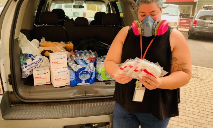 A woman wearing a mask holds hygiene supplies in front of a car full of water bottles and masks.