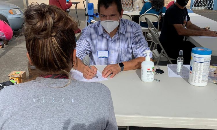 A man in a blue shirt writes something on a piece of paper while talking to a woman with brown hair.