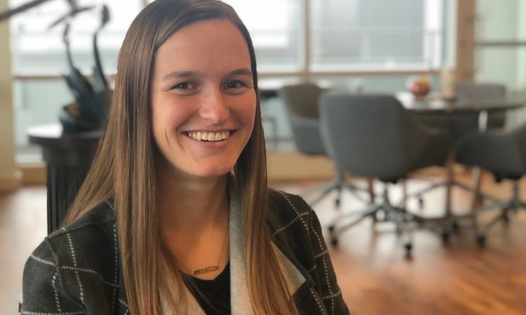 A woman with straight brown hair smiles for the camera while sitting in an office.