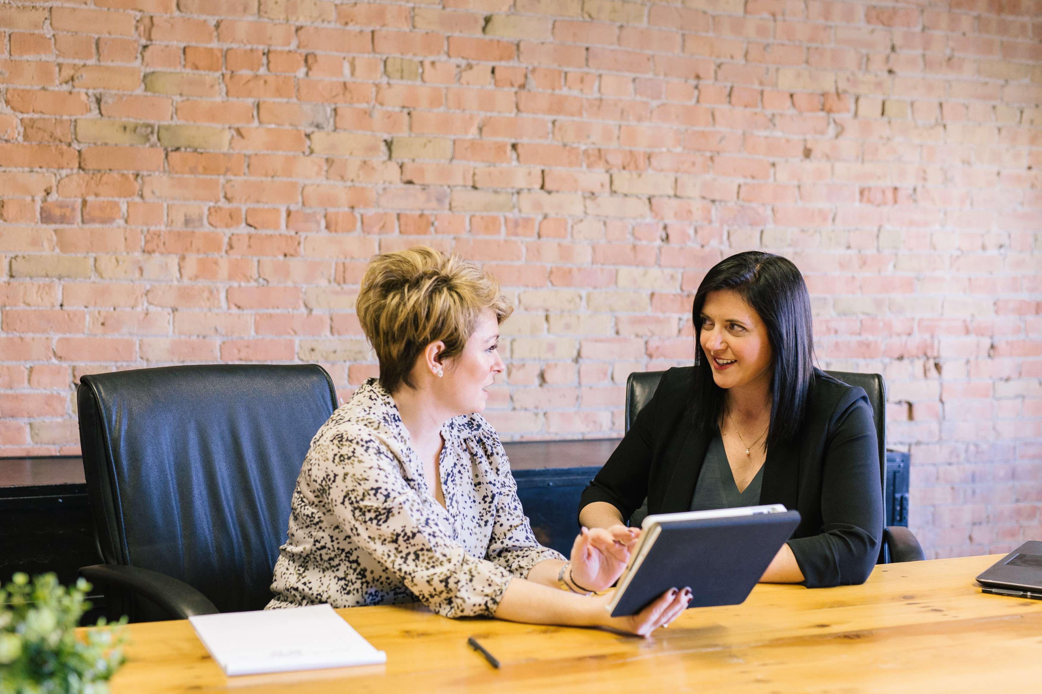 Two woman sit at a table and talk while one holds an iPad.