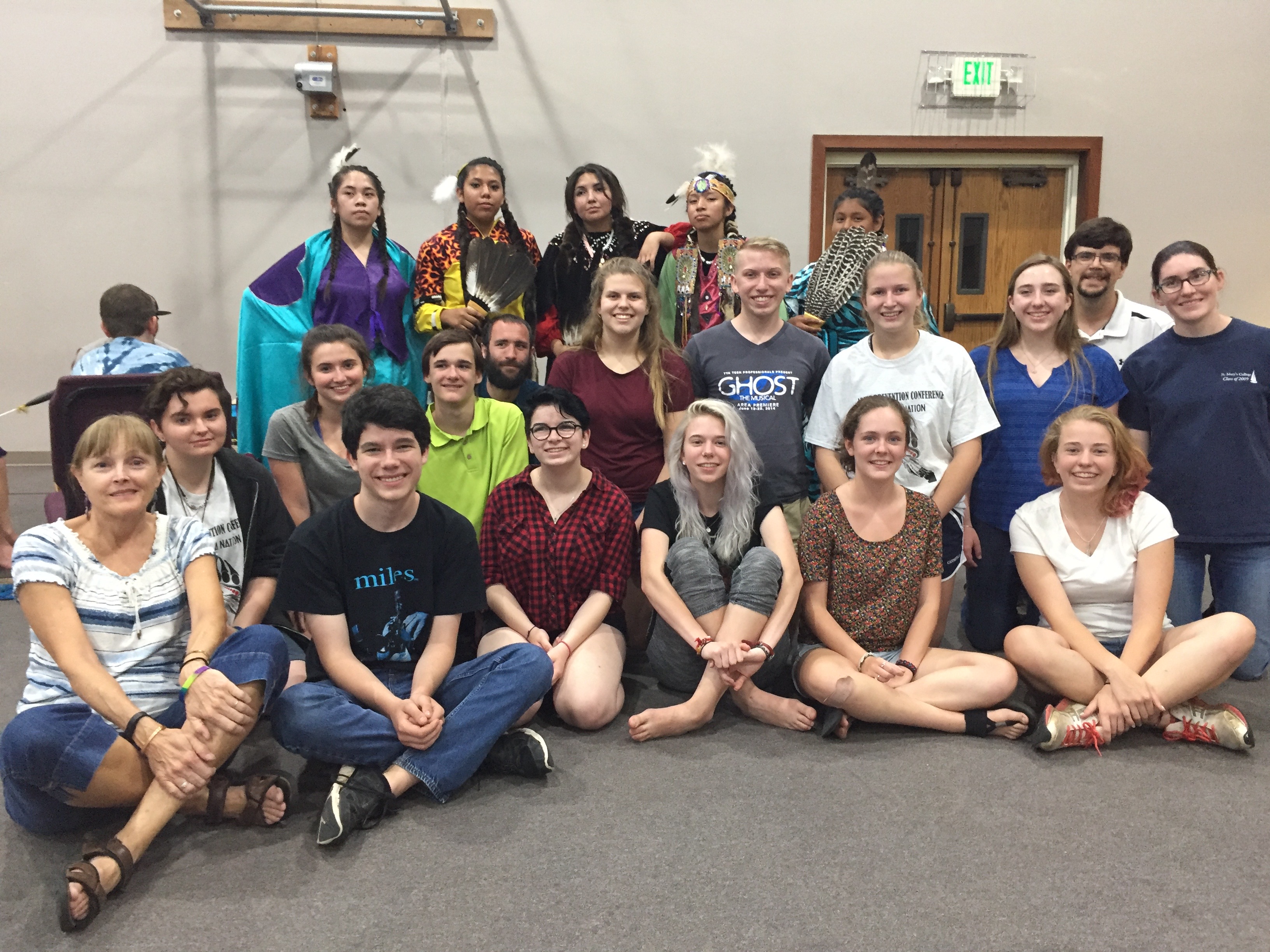 Twenty-one people, some of them wearing traditional Yakama clothing, smile for a picture inside a room