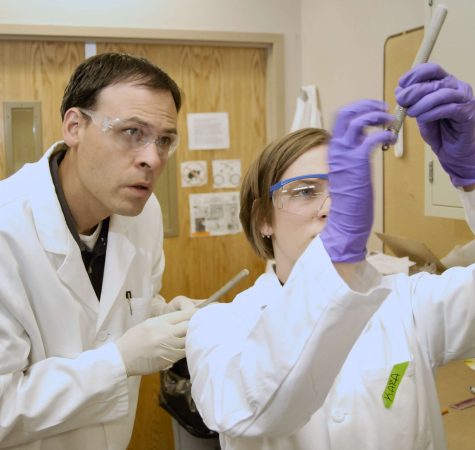 A man with short dark hair wearing a white lab coat and protective goggles and a woman with straight brown hair wearing a white lab coat, protective goggles, and purple gloves observe something in a lab.