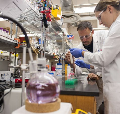 A man with short dark hair wearing a white lab coat and protective goggles and a woman wearing a white lab coat, protective goggles, and purple gloves conduct an experiment in a lab.