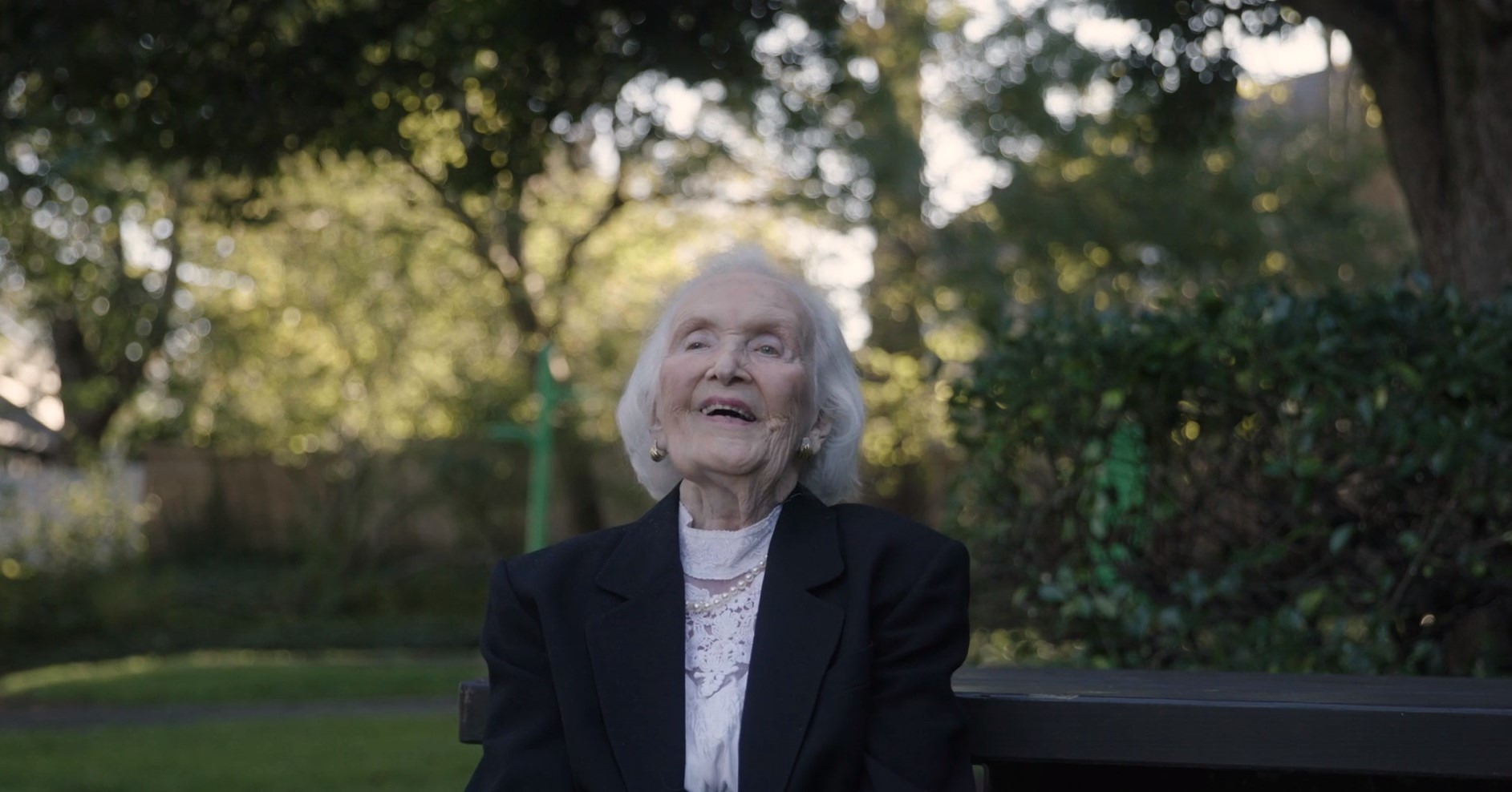 A woman with short white hair wearing a black blazer, a white shirt, pearl necklace, and pearl earrings laughs while sitting on a park bench with trees in the background.