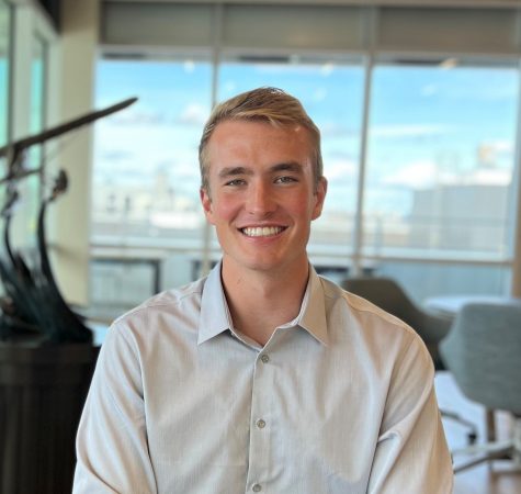 A man with blond hair wearing a light blue button-down shirt smiles at the camera in an office setting.