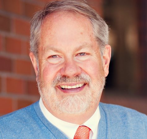 A man with gray hair and facial hear wearing a white shirt, blue sweater, and red tie smiles for the camera.